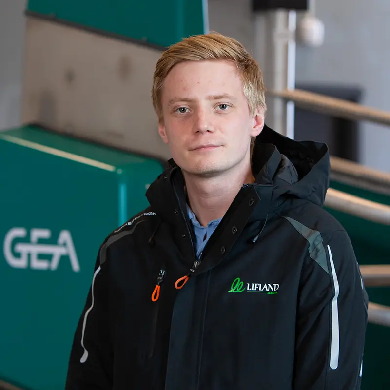 En person med kort blondt hår iført svart jakke står foran grønne Lifland-maskiner med «GEA» synlig på.