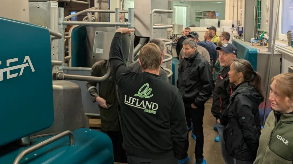 En gruppe mennesker i et fjøs observerer en demonstrasjon av en person iført en "Lifland"-jakke i nærheten av Melkerobot.