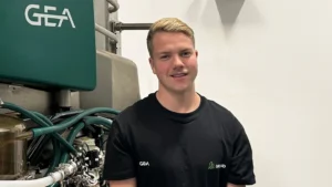 En person i svart GEA-skjorte står foran industrielt utstyr med grønne slanger og GEA-logo, ved siden av en Melkerobot.