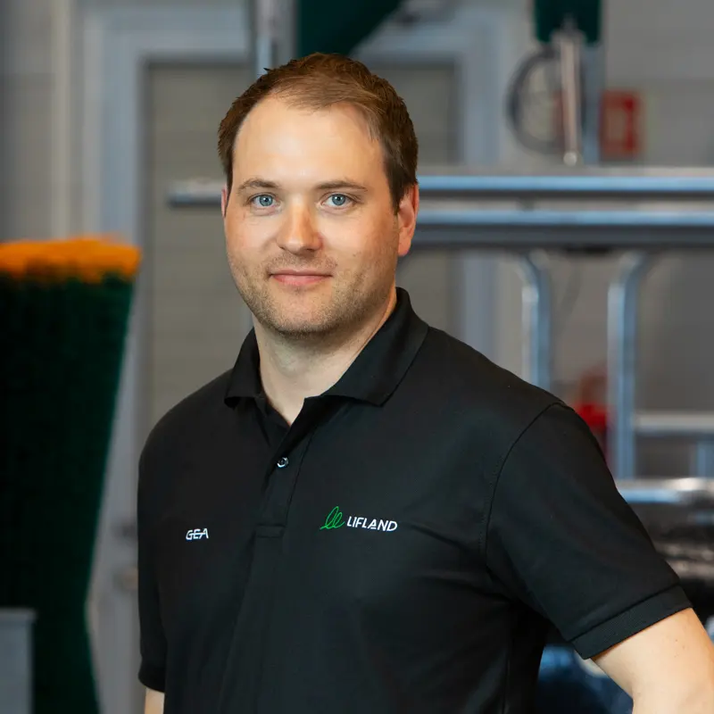 En mann i svart "Lifland"-merket polo står innendørs med industrielt utstyr synlig i bakgrunnen, klar til å hjelpe til med eventuelle Lifland-kontaktforespørsler.