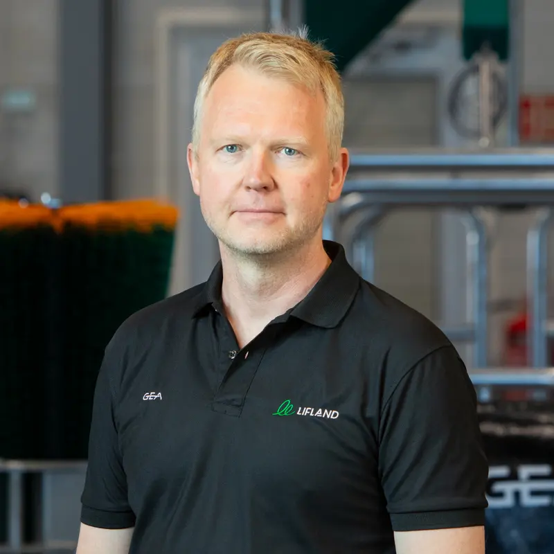 En mann med kort, blondt hår og svart poloskjorte står innendørs foran industrielt utstyr og ser inn i kameraet - muligens på et av Lifland Kontakts anlegg.