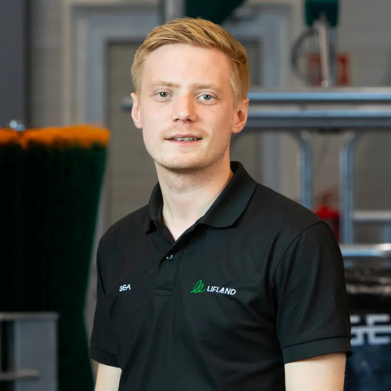 En ung mann med kort, blondt hår og svart Lifland-polotrøye står innendørs og ser inn i kameraet - kanskje klar til å hjelpe deg med eventuelle Lifland-kontaktbehov.