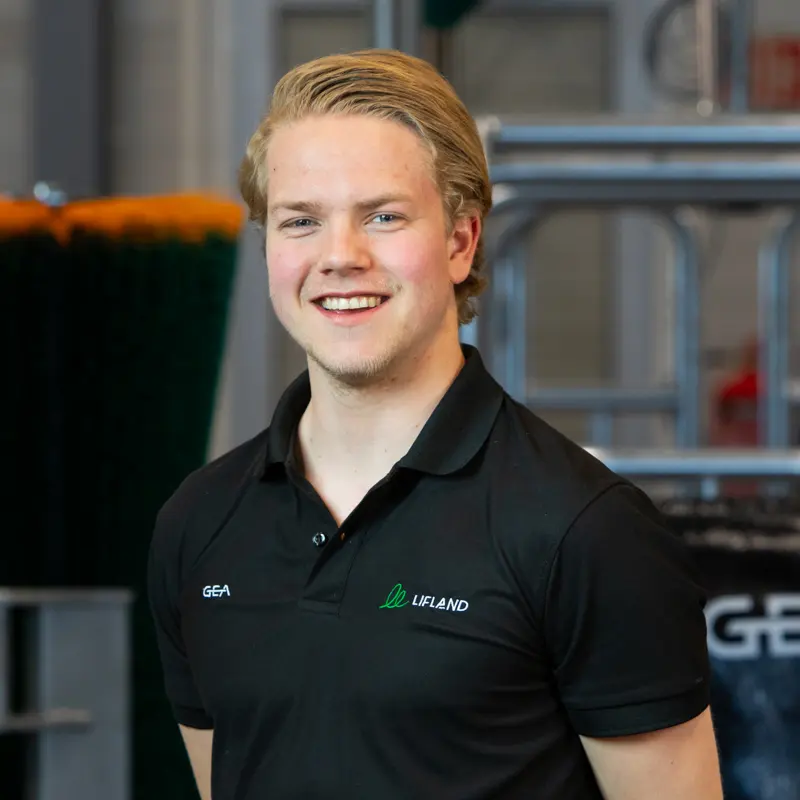 En ung mann med blondt hår smiler til kameraet, iført svart polo med "GEA"- og "Lifland"-logo, står innendørs med industrielt utstyr i bakgrunnen. For mer info, se Lifland kontaktdetaljer.