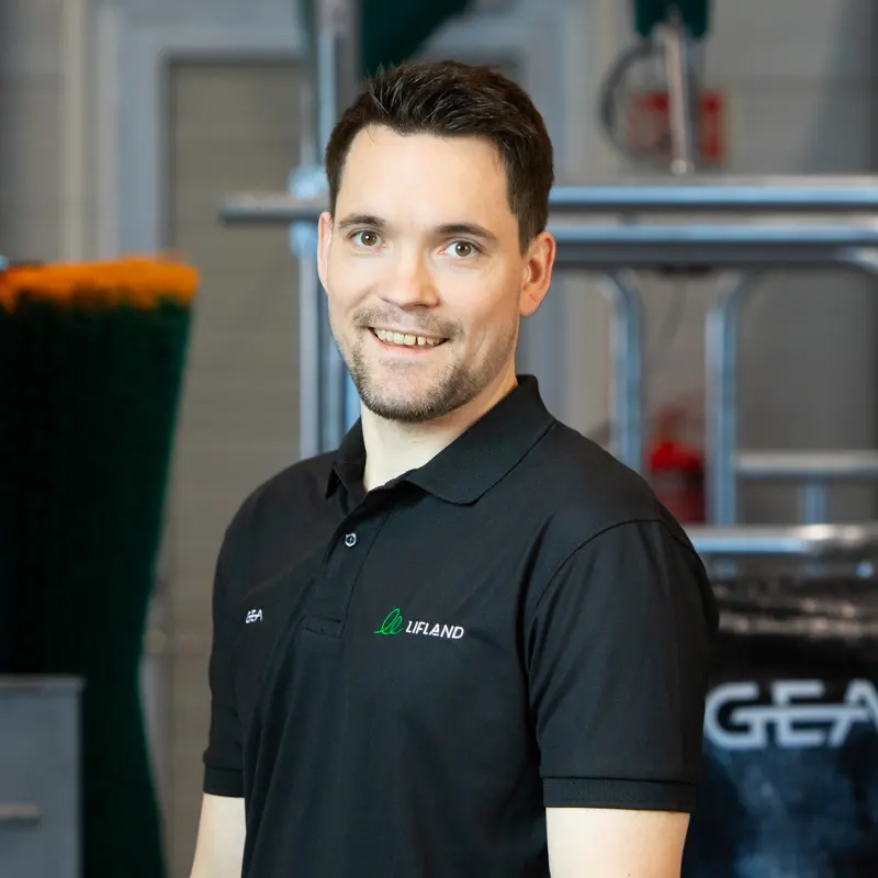 En mann med kort, mørkt hår, iført svart polo med logoene "GEA" og "Lifland Kontakt", står innendørs med industrielt utstyr i bakgrunnen.