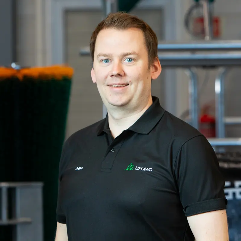 En mann i svart polo med "Lifland"-logo står innendørs og ser inn i kameraet. Industrielt utstyr er synlig i bakgrunnen. For mer informasjon, se Lifland kontakt.