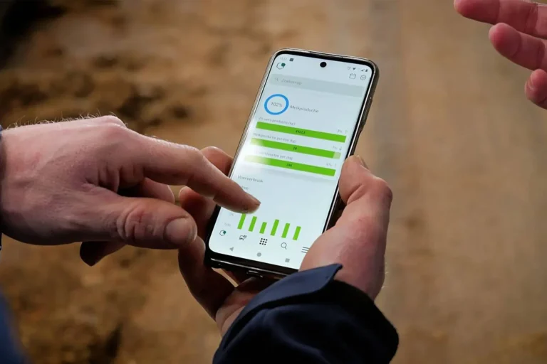 En person holder en smarttelefon som viser Melkerobot-statistikkappen med fremdriftslinjer og grafer, mens en annen hånd peker på skjermen.