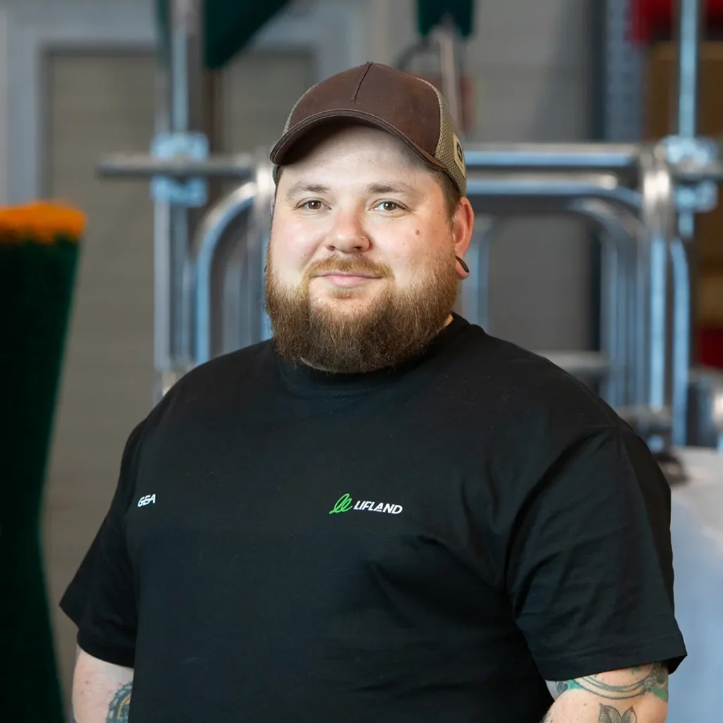 En mann med skjegg, iført svart t-skjorte og brun caps, står innendørs foran industrielt utstyr og ser inn i kameraet - muligens klar for en lifland kontakt.