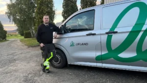 En mann i arbeidsklær, Thomas Aurstad Breiset – ny servicetekniker hos Lifland – står ved siden av en sølvfarget Lifland-varebil som står parkert på en grusvei, med trær og gress i bakgrunnen.