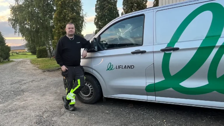 En mann i arbeidsklær, Thomas Aurstad Breiset – ny servicetekniker hos Lifland – står ved siden av en sølvfarget Lifland-varebil som står parkert på en grusvei, med trær og gress i bakgrunnen.